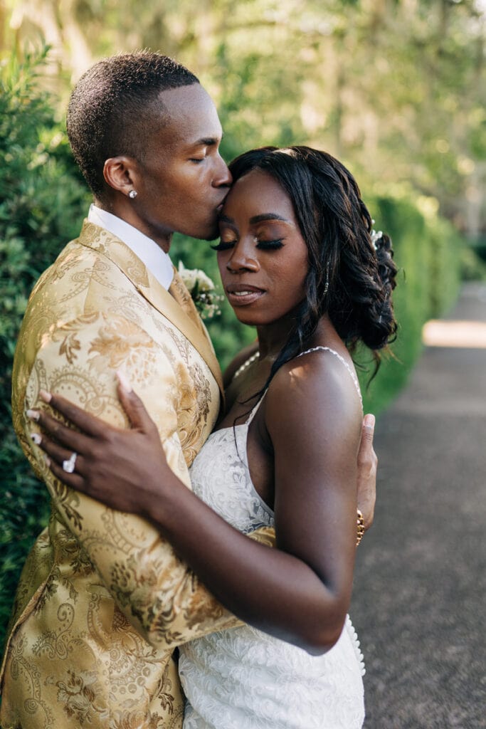 Wedding Photographer, a groom kisses his bride on the head as they embrace outside. He wears a gold colored patterned tux, she wears her wedding dress